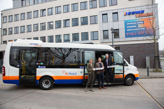 WIMobil startet in Wiesbaden-Biebrich Ein Shuttle-Bus, in dem ein Mann steht. Vor dem Bus stehen zwei weitere Personen, die ein Plakat in den Händen halten