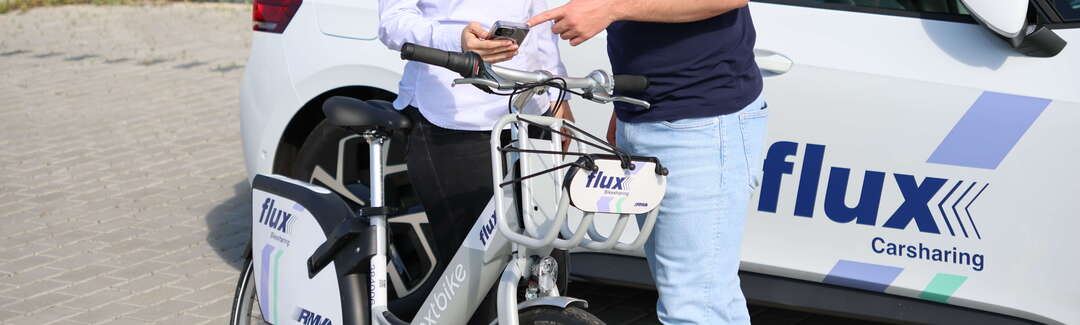 Ein Mann und eine Frau schauen auf ein Handy. Sie stehen vor einem Auto mit der Aufschrift fluxx und einem Rad