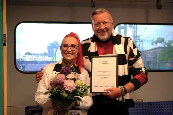 RMV-Lieblingsbusfahrerin 2025: Justyna Cielecka-Drozdz aus Hanau Ein Mann hält eine Urkunde in der Hand, eine Frau steht daneben mit einem Blumenstrauß.