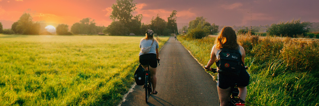 Komoot Radler Sonnenuntergang Radler fahren neben einer Wiese dem Sonnenuntergang entgegen.