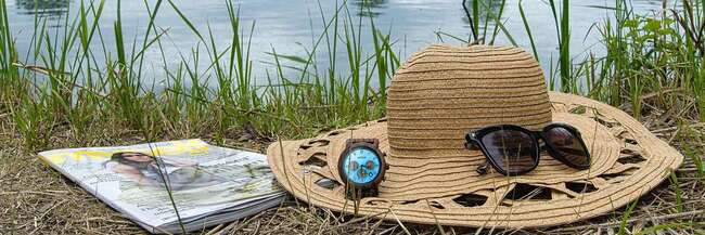 Strohhut, Sonnenbrille, Armbanduhr und Zeitschrift im Gras vor Badesee