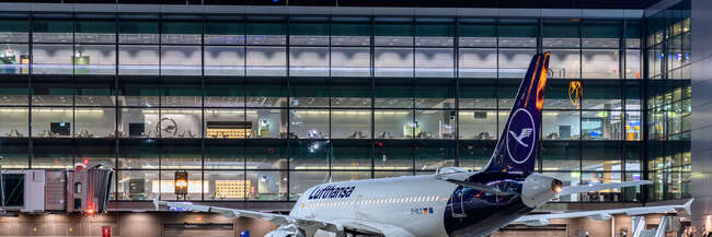 Flughafenterminal mit blauer Neonschrift Frankfurt Airport bei Nacht. Davor steht eine Flugzeug von Lufthansa.