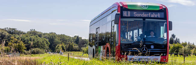 Elektrobus HLB Ein Elektrobus der MTV fährt auf einer Landstraße durch eine grüne Landschaft im Main-Taunus-Kreis. Im Vordergrund sind Wiesenblumen zu sehen, im Hintergrund Bäume unter blauem Himmel.