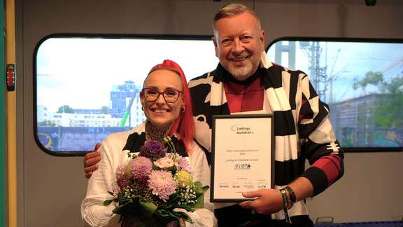 RMV-Lieblingsbusfahrerin 2025: Justyna Cielecka-Drozdz aus Hanau Ein Mann hält eine Urkunde in der Hand, eine Frau steht daneben mit einem Blumenstrauß.