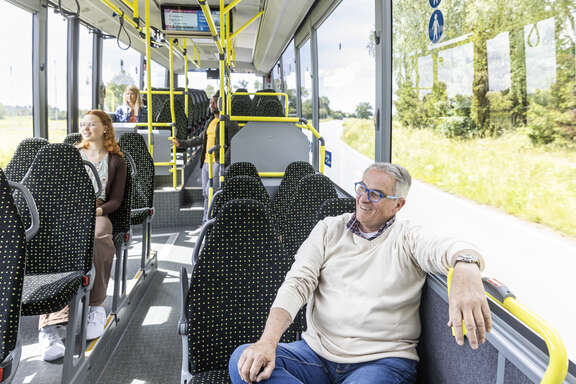 Vier Personen sind im Bus, zwei junge Frauen sitzen auf der linken Seite, auf der rechten Seite sitzt vorne ein Mann und hinten steht ein Mann.